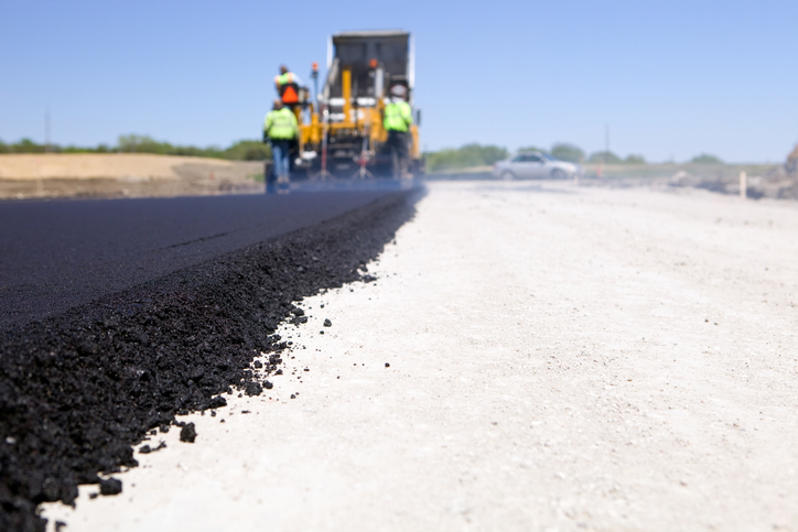 A dump truck is unloading fresh asphalt into a slipform paver, at a new road construction site. Steam is rising from the hot surface directly behind the paver and a car is passing on the cross street. This image could be used equally well for a new driveway project. http://www.banksphotos.com/LightboxBanners/RoadConstruction.jpg